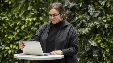 Silmälasipäinen nainen avaa kannettavan tietokoneen pyöreällä, vaalealla pystypöydällä viherseinän edustalla.