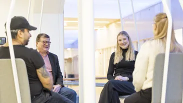 Neljä henkilöä istuvat vastakkain hyvin valaistussa tilassa käytävän varrella ja hymyilevät toisilleen.