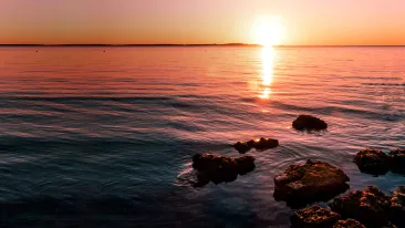 Aurinko laskee oranssin sävyissä taivaanrantaan heijastuen tyynestä vedestä, etualalla rannassa useampi kivi.