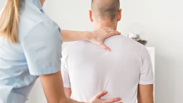 Sisätiloissa sinisessä asussa oleva fysioterapeutti hoitaa tai tutkii asiakasta valkoisessa t-paidassa. Fysioterapeutin kädet ovat asiakkaan yläselän ja olkapään alueella.