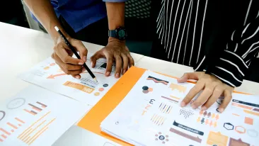 Henkilö kirjoittaa pöydällä olevalle paperille, jota vieressä oleva henkilö raidallisessa kauluspaidassa seuraa.