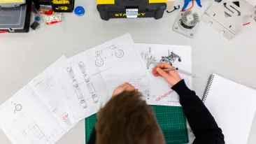 Pöydän ääressä istuva henkilö tarkastelee ja merkitsee insinöörin piirustuksia pöydällä, työkalujen ja mekaanisten osien äärellä.