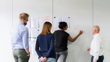Neljän henkilön ryhmä tekee yhteistyötä valkotaulun äärellä. Yksi henkilö piirtää tussilla taululle, johon on kiinnetty papereita.