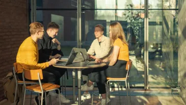 Neljä nuorta istuvat valoisassa tilassa kannettavien tietokoneiden äärellä pyöreässä pöydässä.