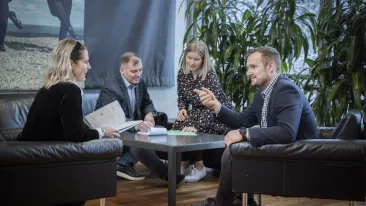 Neljä henkilöä istuvat viherkasvien reunustamalla sohvaryhmällä ja keskustelevat hyvässä hengessä toistensa kanssa.