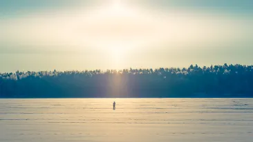 Aurinko paistaa kirkkaalta taivaalta talvipäivänä, lumipeitteisen järven jäälle on pysähtynyt yksinäinen sauvakävelijä.