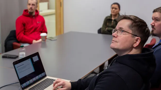 Ihmiset katsovat neuvotteluhuoneen pöydän äärellä näyttöä kohti, pöydällä on avoin kannettava tietokone.