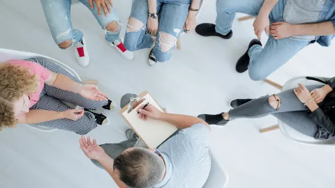 Ryhmä ihmisiä, nuoria ja aikuisia, on kokoontunut ympyrään keskustelemaan. Yksi henkilö ottaa muistiinpanoja lehtiöön.