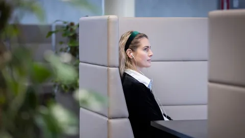 Vaaleahiuksinen nainen istuu ajatuksissaan huonekasvien ympäröimällä sohvaryhmällä ja kuuntelee jotakin nappikuulokkeilla.