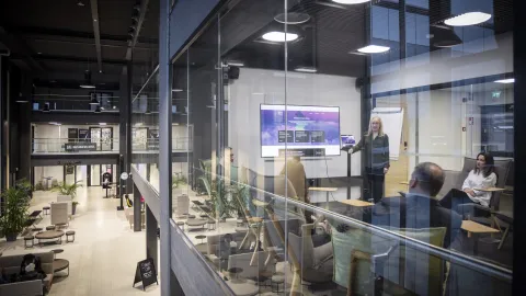 Korkeassa avoimessa tilassa olevan neuvotteluhuoneen lasien läpi näkyy joukko ihmisiä, joista yksi pitää esitystä ja muut kuuntelevat keskittyneesti.