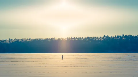 Aurinko paistaa kirkkaalta taivaalta talvipäivänä, lumipeitteisen järven jäälle on pysähtynyt yksinäinen sauvakävelijä.