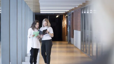 Kaksi henkilöä seisovat vierekkäin valoisalla käytävällä ja keskittyvät tutkimaan muistiinpanokirjaa.