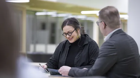 Kaksi henkilöä istuvat vierekkäin, toinen selkä kameraan päin, ja tutkivat papereita pöydän äärellä.